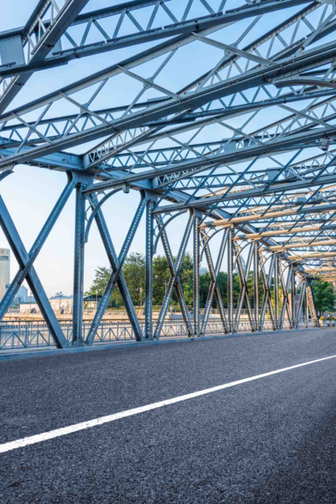 A road runs beneath a metal beam bridge, illustrating the connection between two sides and the strength of its construction.